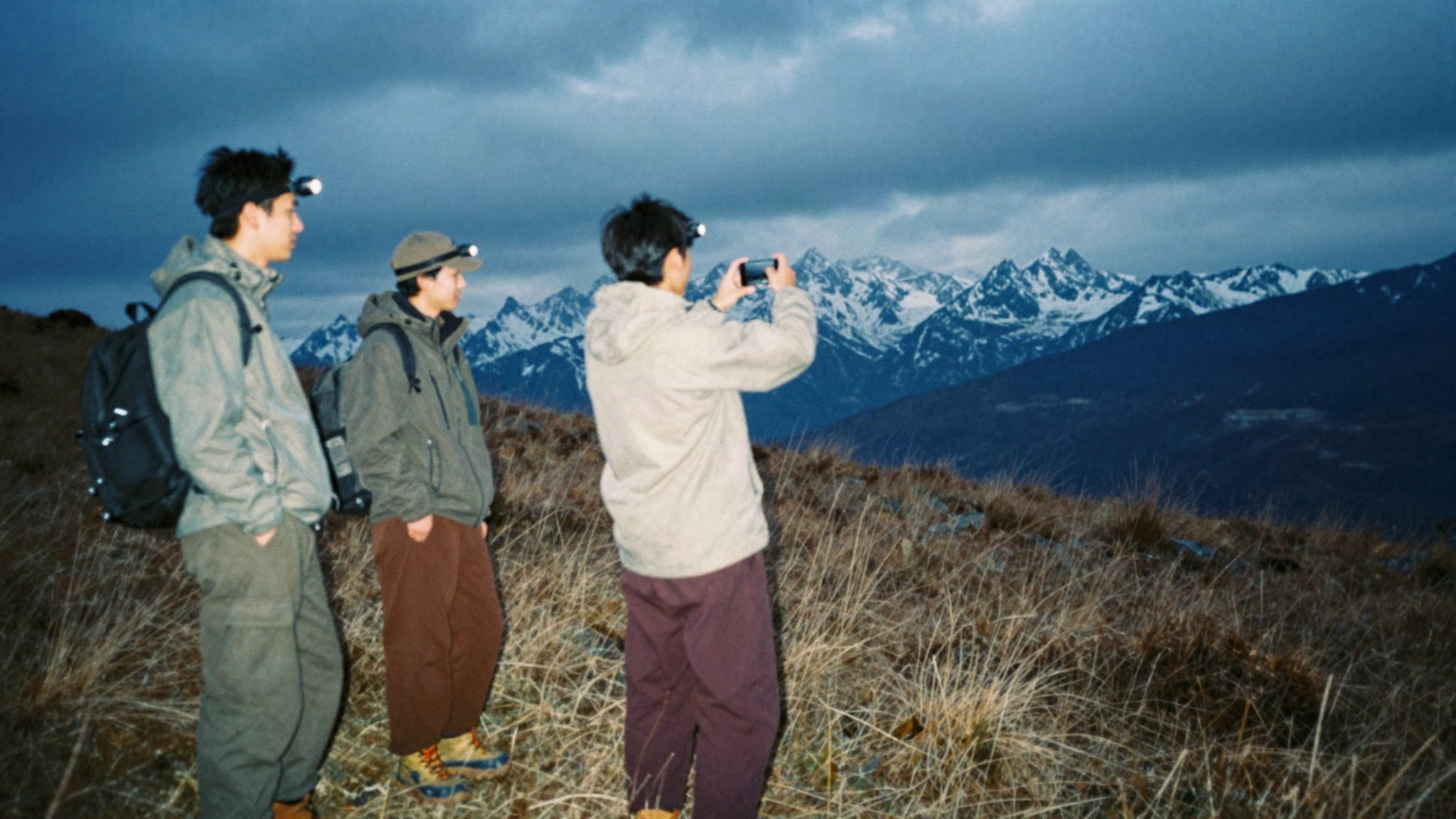 Three people in head torches on a hillside before sunrise, one framing the mountains on a phone.
