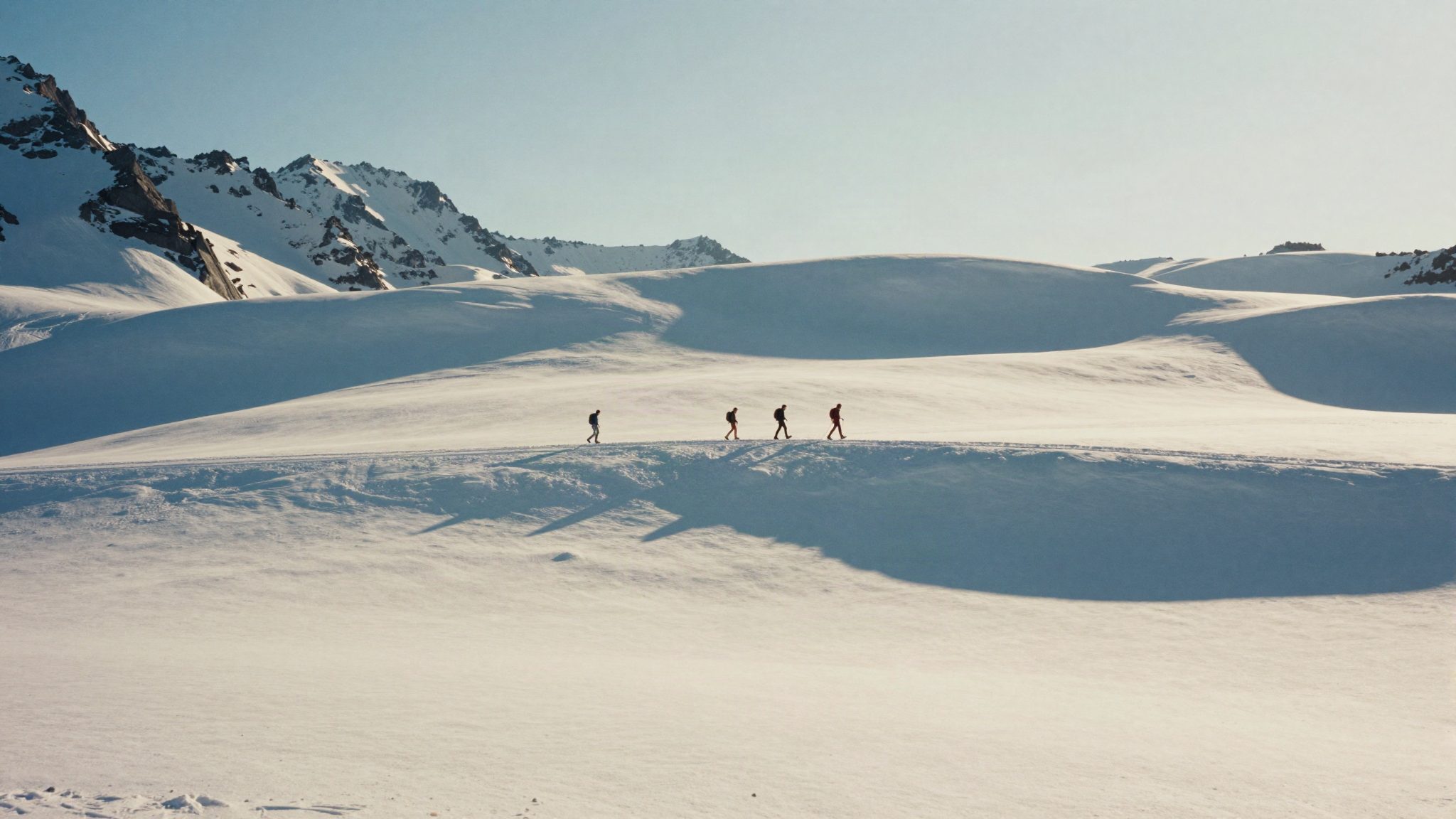 Four people walking in single file across a sunlit glacier, their shadows long on the snow.